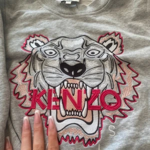 Grå Kenzo sweatshirt med tiger - Säljer en grå sweatshirt från Kenzo med ikonisk broderad tiger och röd Kenzo-logga på bröstet. Tröjan har långa ärmar, rund hals och coola detaljer i rosa och svart. Perfekt för dig som gillar streetstyle och vill sticka ut med ett snyggt statement-plagg.