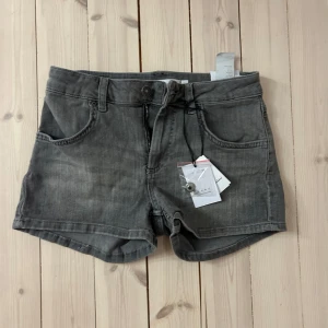 Grå lågmidjade jeansshorts Subdued - Snygga grå jeansshorts från Subdued med låg midja och klassisk femficksdesign. Shortsen har knapp och dragkedja framtill, bälteshällor och coola bakfickor med broderad detalj. Dessa är endast testade, och har fortfarande kvar lappar och extra knappen. Köpta i London, säljer då de ej kommer till användning, de är i storlek 28 🫶