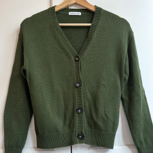 Grön kofta från Designers Remix - Snygg grön kofta från Designers Remix med v-ringad hals och svarta knappar framtill. Den är stickad i mjukt material och har långa ärmar samt ribbade muddar vid ärmslut och nederkant. Perfekt att slänga på sig när det är lite kyligt ute.