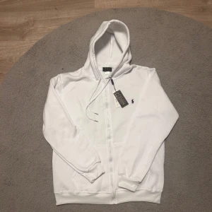 Vit hoodie från Polo Ralph Lauren - Säljer en clean vit hoodie från Polo Ralph Lauren i storlek L. Tröjan har huva med snörning, dragkedja framtill och en stor ficka på magen. Klassisk Polo-logga broderad på bröstet. Perfekt för chill dagar och enkel att matcha med jeans eller joggers.Höjden är 65 cm och Breden är 50 cm.Den är helt ny med tagg.💬Skriv gärna om du har frågor! 📦 Skickas inom 24 timmar! 📉 Kan gå ner lite i pris vid snabb affär! 🕒 Aktiv nästan hela tiden svarar snabbt! 