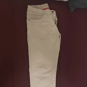 Beige chinos från Tommy Hilfiger - Snygga beige chinos från Tommy Hilfiger med klassisk femficksdesign och knappgylf. Byxorna har en rak passform och är tillverkade i mjuk bomull som är skön att bära. Perfekta för dig som gillar stilrena och enkla byxor med diskreta detaljer.