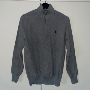 Grå stickad tröja från Ralph Lauren - Snygg grå stickad tröja från Ralph Lauren med half zip och hög krage. Tröjan har långärmade ribbade muddar och en liten marinblå logga broderad på bröstet. Perfekt för dig som gillar klassisk stil med en modern touch.