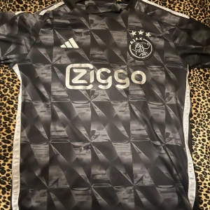 Ajax svart matchtröja Adidas - Ajax fotbollströja från Adidas i svart med geometriskt mönster och silverfärgade detaljer. Kortärmad med Ziggo sponsortryck och Ajax klubbmärke på bröstet. Materialet är lätt och andas, perfekt för träning eller match. Snygga vita ränder längs sidorna.