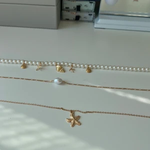 Pärlarmband och halsband med guldberlocker - Säljer ett pärlarmband med små guldiga berlocker i form av snäckor och sjöstjärnor, samt två tunna guldfärgade halsband, ett med en vit pärla och ett med en sjöstjärna som hängsmycke. Perfekt för dig som gillar strandvibbar och detaljer.