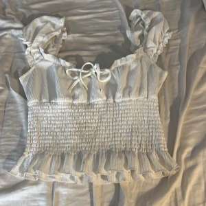 Vit volangblus från Gina Tricot - Supersöt vit blus från Gina Tricot med volanger och smockad midja. Blusen har korta puffärmar, knyt framtill och ett lätt randigt mönster. Perfekt för sommarens alla häng! Materialet känns som mjuk bomull och toppen har en feminin v-ringad form.