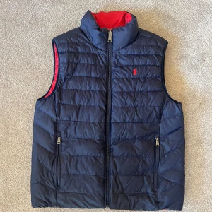 Dunväst från Polo Ralph Lauren, vändbar - Vändbar dunväst från Polo Ralph Lauren i storlek L. Ena sidan är marinblå med röd logga, andra sidan är röd med marinblå logga. Västen har ståkrage, två fickor med dragkedja och quiltad design. Materialet är återvunnen nylon och polyester.
