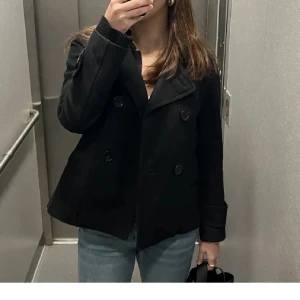 Svart skepparkavaj från H&M - Säljer en svart skepparkavaj från H&M med dubbelknäppning och bred krage. Jackan har långa ärmar med snygga knappdetaljer vid mudden och en klassisk rak passform. Perfekt för dig som gillar stilrena och tidlösa plagg.