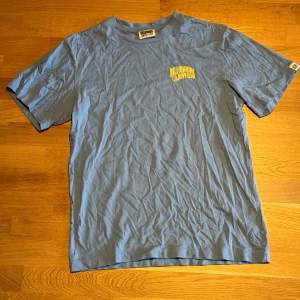 Blå Billionaire Boys Club t-shirt - Snygg blå t-shirt från Billionaire Boys Club med gul logga och astronauttryck på bröstet. Klassisk rund hals och korta ärmar. Mjuk bomull som känns skön mot huden. Perfekt för dig som gillar streetwear och vill sticka ut med en cool detalj på ryggen.