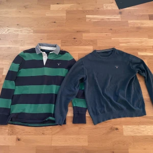 GANT randig pikétröja & blå tröja - Två tröjor från GANT i storlek S och M. Ena är en pikétröja med breda gröna och mörkblå ränder, vit knappslå och grå krage. Den andra är en  mörkblå tröja med rund hals och broderad liten  GANT-logga på bröstet. Den randiga saknar 2 knappar. Den mörklblåa är storlek M men passar storlek S.