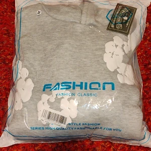 Grå hoodie med vita blommor - Grå hoodie i storlek M med vita blommiga tryck över hela tröjan. Tröjan har huva, magficka och långa ärmar. Perfekt för dig som gillar chill och snygg streetstyle. Materialet känns mjukt och skönt, troligen bomull eller polyester.