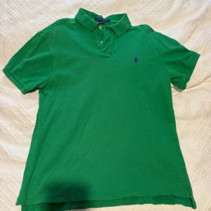 Grön Polo Ralph Lauren t-shirt L - Snygg grön t-shirt från Polo Ralph Lauren i slim fit-modell. Klassisk pikékrage med två knappar och lila broderad logga på bröstet. Tillverkad i mjuk bomull som är skön mot huden. Perfekt för dig som gillar stilren och sportig look.