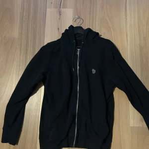 Svart hoodie med dragkedja och huva - Svart hoodie från Paul & Smith med dragkedja framtill och huva med snörning. Liten broderad tigerlogga på bröstet. Tröjan har långa ärmar och ribbade muddar. Perfekt för chill dagar och streetwear-stil.