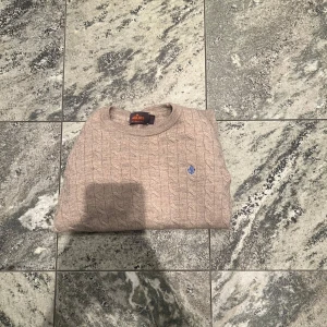 Beige kabelstickad tröja Morris XS - Snygg beige kabelstickad tröja från Morris i 100% merinoull. Rund hals, långärmad och med ett litet blått broderat emblem på bröstet. Perfekt för dig som gillar klassisk stil med en modern touch. Supermjuk och skön att ha på sig.