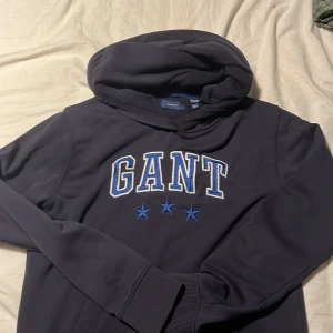 Mörkblå hoodie från GANT med tryck - Snygg mörkblå hoodie från GANT med stort broderat logotryck och tre blå stjärnor på bröstet. Tröjan har huva, långa ärmar och ribbade muddar. Perfekt för chill dagar och streetstyle. Materialet är mjukt och skönt, troligtvis bomull eller polyester.