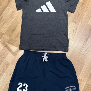 Svarta Adidas shorts med röd logga - Svarta shorts från Adidas med röd Adidas-logga på ena benet och liten vit logga på andra sidan. De har elastisk midja med röd snörning och är gjorda i lätt syntetmaterial, perfekt för sport eller chill. Snygg och enkel design som funkar till det mesta. Även en grå Adidas T-shirt i fint skick med ett par shorts från Vinson i fint skick. 