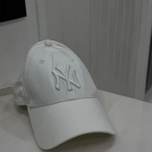 Vit New Era Yankees keps 9FORTY - Snygg vit keps från New Era med klassiska NY Yankees-loggan broderad framtill. Modellen är 9FORTY med böjd skärm och justerbart spänne bak i metall. Kepsen är tillverkad i 100% bomull och har diskreta broderade detaljer på sidan.