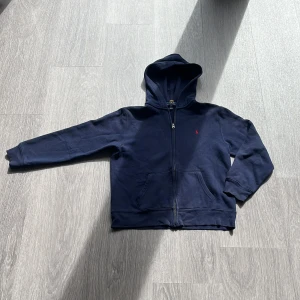 Marinblå ziphoodie från Polo Ralph Lauren - Snygg marinblå hoodie från Polo Ralph Lauren med dragkedja och huva. Tröjan har ett litet rött Polo-logo broderat på bröstet, två fickor fram och ribbade muddar vid ärmslut och nederkant. Perfekt för chill dagar och enkel att matcha med jeans. Påse medföljer men har defekter. Hör av er vid minsta funderingar🤗