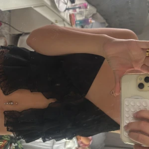 Svart spetsig croppad korsett-topp - Säljer en svart croppad korsett-topp med spetsdetaljer och volanger. Toppen har en v-ringning, knappar framtill och är ärmlös med tunna axelband. Perfekt för dig som vill ha en edgy och trendig look.