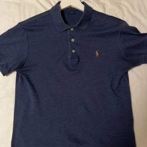 Marinblå pikétröja från Ralph Lauren - Snygg marinblå pikétröja från Ralph Lauren med klassisk krage och tre knappar framtill. Slim fit-modell med korta ärmar och ikoniska broderade polospelaren på bröstet. Perfekt för dig som gillar stilren och enkel design. Säljer denna för jag har växt ur den! 