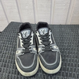 Louis Vuitton svarta sneakers #54 - Svarta och grå sneakers från Louis Vuitton med coola LV-loggor på plösen och sidan, samt #54 tryckt bak på hälen. Skorna har snörning och en platt sula. Materialet är en mix av skinn och syntet, med detaljer i vitt och grått. Perfekt för dig som gillar streetstyle och exklusiva märken. Storlek 43 men passar 44.