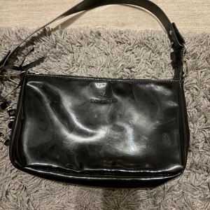 Svart axelväska från SIXTH SENSE - Snygg svart axelväska från SIXTH SENSE i glansigt skinnmaterial. Väskan har en bred axelrem med metallspänne och dekorativa detaljer vid dragkedjan. Perfekt storlek för att få plats med det viktigaste och passar till många olika stilar.