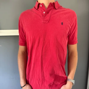 Röd Ralph Lauren piké  - Säljer en klassisk röd Polo t-shirt från Ralph Lauren i storlek M. T-shirten har korta ärmar, krage med knappar och den ikoniska broderade Polo-loggan på bröstet. Tillverkad i 100% bomull för skön och luftig känsla. Perfekt för dig som gillar stilren och tidlös design.