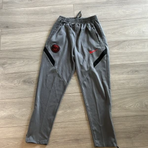 Grå PSG x Nike träningsbyxor - Grå träningsbyxor från Nike med Paris Saint-Germain-logga i rött och svart på ena benet och röd Nike Swoosh på andra. Byxorna har svarta detaljer, elastisk midja med snörning och dragkedja vid benslut. Perfekta för fotboll eller chill.