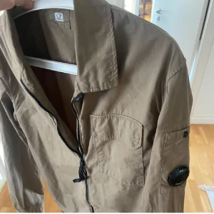 CP Company brun overshirt med zip storlek M - Snygg brun overshirt från CP Company med klassisk krage, bröstficka och ikonisk linsdetalj på ärmen. Storlek M men skulle säga att den sitter mer som S. Bara att höra av sig vid frågor!