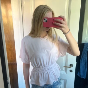 Ljusrosa blus med resår i midjan - Säljer en ljusrosa blus med korta ärmar och rund hals. Toppen har en bred resår i midjan som ger en snygg volang-effekt nedtill. Perfekt till jeans eller kjol för en chill och söt look. Materialet känns mjukt och lätt.