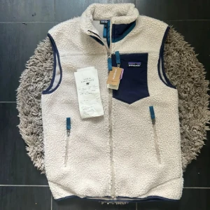 Vit fleeceväst från Patagonia S - Säljer en sjukt snygg vit fleeceväst från Patagonia i storlek S. Västen har en hög krage, dragkedja framtill och två sidofickor med blå detaljer. Bröstfickan är marinblå med Patagonia-logga. Perfekt för lager-på-lager och chill stil.