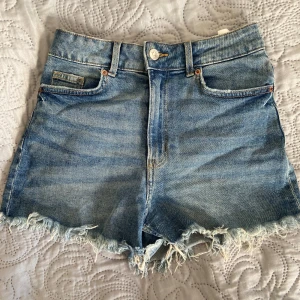 Blå jeansshorts med fransig kant -  Blå jeansshorts med hög midja och fransig kant längst ner. Knapp och dragkedja framtill. Kontakta vid funderingar 💗