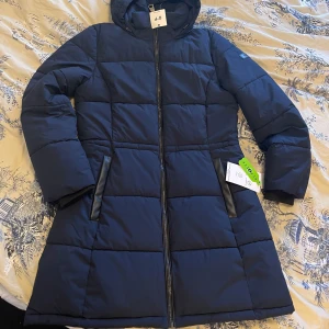 Marinblå pufferjacka från DKNY - Snygg marinblå pufferjacka från DKNY med huva och raka, quiltade paneler. Jackan har dragkedja framtill, två fickor med svarta detaljer och en diskret DKNY-logga på ärmen. Materialet är polyester och jackan är vattenavvisande med stretch. Perfekt för kalla dagar.helt ny