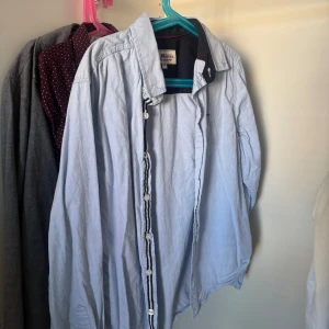 Ljusblå skjorta Hampton Republic 152 - Snygg ljusblå skjorta från Hampton Republic med klassisk krage och vita knappar. Skjortan har mörkblå detaljer på insidan av kragen och knappslån, vilket ger en cool kontrast. Perfekt för dig som gillar stilrena och tidlösa plagg.