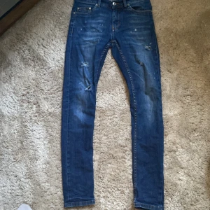 Blå jeans med slitningar - Säljer ett par blå jeans med coola slitningar och lätt tvättad look. Jeansen har klassisk femficksdesign, smal passform och är tillverkade i stretchigt denim för skön känsla. Perfekta för dig som gillar en streetig stil och vill ha jeans med attityd.