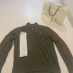 Olivgrön half zip tröja Morris S - Snygg olivgrön stickad tröja från Morris med half zip och ribbad krage. Tröjan har ett litet broderat emblem på bröstet och ribbade muddar vid ärmslut och nederkant. Perfekt för dig som gillar klassisk stil med en modern touch. Tröjan är i jättebra skick och har inga defekter. Kvittot till tröjan finns. Nypris är 1700kr. Johnelles påsen kommer med på köpet och de gör även kvittot. Hör av er vid minsta fundering