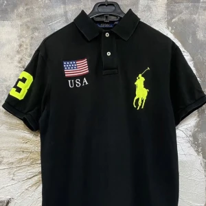 Svart Rare Ralph Lauren polo med USA-tryck - Svart kortärmad  polo från Ralph Lauren med neongult broderat polospelare-logo, stor siffra 3 på ärm och rygg, samt USA-flagga. Inga fläckar som ny.
