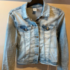 Ljusblå jeansjacka från &Denim - Snygg ljusblå jeansjacka från &Denim med klassisk krage, silverfärgade knappar och två detaljfickor. Jackan har långa ärmar och är tillverkad i jeansmaterial. Använd några gånger, gammal modell. 