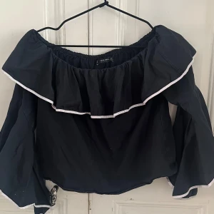 Svart offshoulder blus från Zara - Supersnygg svart offshoulder blus från Zara med bred volang runt halsen och vita kanter. Ärmarna är långa och vida med samma vita kantdetalj. Perfekt för dig som gillar statement-plagg med cool siluett och romantisk vibe.