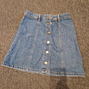 Blå jeanskjol med knappar framtill - Säljer en klassisk blå jeanskjol med A-linjeform och knappar längs hela framsidan. Kjolen har hällor för bälte och är perfekt för dig som gillar en enkel men snygg denimlook. Passar dig som vill ha en kortkjol med retrovibbar.