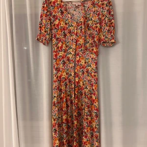 Blommig långklänning från ONLY - Superfin långklänning från ONLY med färgglatt blommigt mönster i rött, rosa, gult och grönt. Klänningen har korta puffärmar, v-ringning med knytband och markerad midja. Perfekt för sommarens alla tillfällen och ger en härlig boho-vibe.