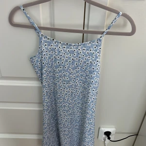 Blommig blå kortklänning 1st New - Söt blå kortklänning från 1st New med vita och svarta blommor. Klänningen har smala axelband och är ärmlös, perfekt för varma dagar. Materialet är mjukt och lätt, och den har en avslappnad passform som ger en skön känsla. Superfin till sneakers eller sandaler!