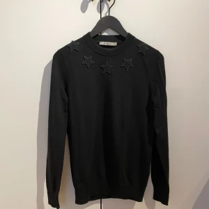Svart stickad tröja Givenchy med stjärnor - Svart stickad tröja från Givenchy med snygga svarta stjärnapplikationer på axlar och runt halsen. Tröjan har rund hals och långa ärmar, perfekt för dig som gillar stilrena detaljer med en twist. Passar dig som vill ha en cool och enkel look.