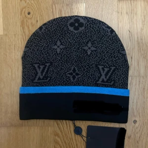 Svart och blå mönstrig mössa  - Svart och blå mössa med ikoniskt monogrammönster. Mössan har en blå kant nedtill som ger en snygg kontrast. Perfekt för dig som vill ha en lyxig och stilren accessoar till vintern.