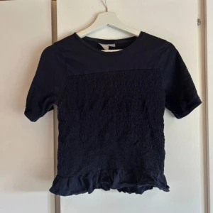 Mörkblå topp med volangkant - En mörkblå topp med korta ärmar och rund hals. Framsidan har smockad struktur och nertill finns en söt volangkant som ger en extra detalj. Toppen är i mjukt bomullsmaterial och har en enkel, stilren look som passar till jeans eller kjol. Storleken är 170 men passar xs/s/m 