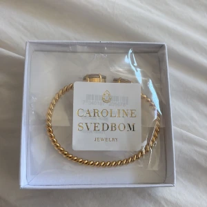 Guldarmband med rosa stenar Caroline Svedbom - Oanvänt!! Snyggt guldarmband från Caroline Svedbom med tvinnad design och två ljusrosa kristaller i ändarna. Perfekt accessoar för att lyfta din outfit med lite extra bling. Kommer i original ask.