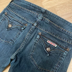 Låga Vintage Hudson bootcut jeans i mörkblå denim - Snygga bootcut jeans från Hudson i mörkblå denim med klassiska kontrastsömmar och dubbla knappar i midjan. Bakfickorna har coola detaljer med knappar och logopatch. Jeansen har låg midja och är tillverkade i mjukt jeansmaterial. | Min mammas gamla jeans från 2000 talet, dom är insydda i midjan, och uppsydda några cm. Därför stämmer inte storleken som står på lappen. Mått : Midja - 37cm Innerben - 77cm (+3) Ytterben - 98cm (+3) Midjehöjd - 20cm