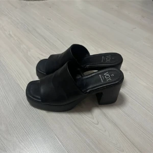 Svarta klackade sandaler från Vox - Svarta sandaler från Vox med bred rem över foten och öppen tå. Snygg chunky klack och platt sula ger en cool vibe. Tillverkade i skinnimitation med stilren design och fyrkantig tå. Perfekta för dig som gillar att sticka ut med enkla men trendiga skor.