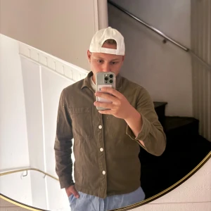 Olivgrön overshirt från East West - Säljer en olivgrön overshirt från East West i storlek M. Skjortan har två bröstfickor, vita knappar och klassisk krage. Materialet är mjuk bomull med lite struktur, perfekt att slänga på över en t-shirt. Snygg och enkel att styla.