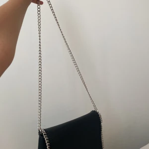 Svart axelväska med kedja - Snygg svart axelväska med silverfärgad kedja som axelrem och detaljer längs kanterna. Väskan har ett stilrent, rektangulärt format och magnetlås. Insidan är rymlig med ett fack, perfekt för mobilen och småsaker. Materialet är slätt och ger en cool vibe. Den har bara andvänts max 4 gånger! Jag säljer den då är för kort i kedjan för mig , den är i jätte fint skick och har inte repor eller nått konstigt på den! Perfekt för att ha mobil och lite andra små saker och annat / 💘väskan är fån Lindex!🩷