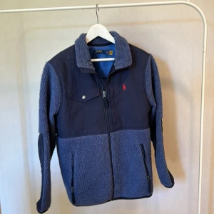 Blå teddyjacka från Polo Ralph Lauren - Snygg blå teddyjacka från Polo Ralph Lauren med mjuk fleece och detaljer i mörkare blått tyg. Jackan har dragkedja framtill, bröstficka med knapp, två sidofickor med dragkedja och klassisk röd logga på bröstet